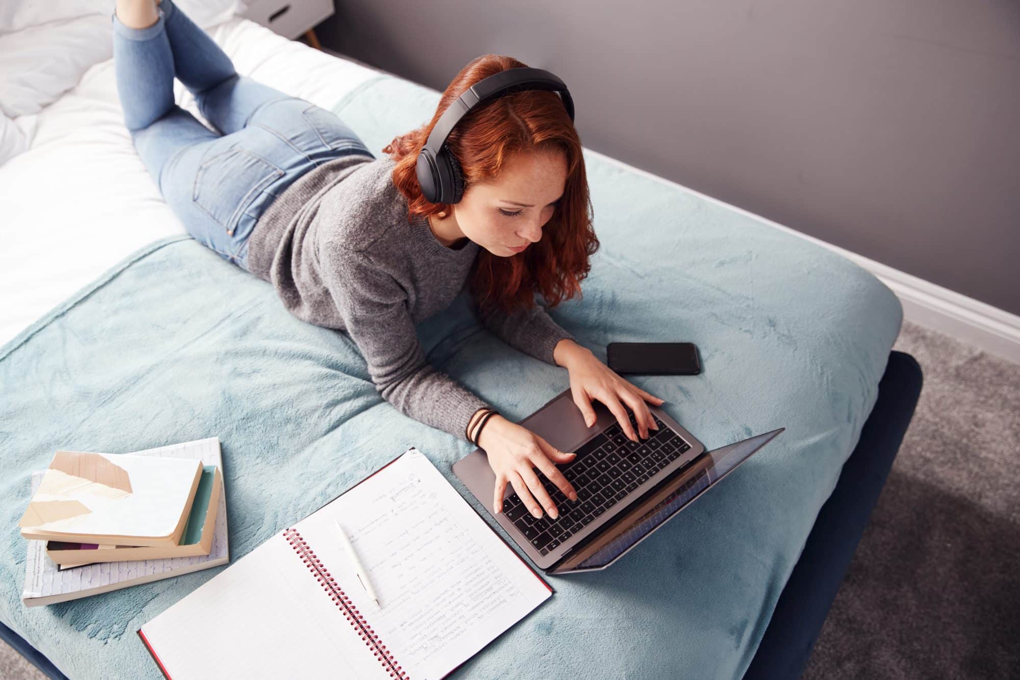 looking down on female college student wearing headphones lying on bed working on laptop